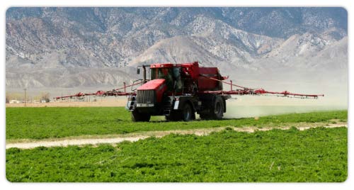 A red agricultural sprayer applying fertilizer across a green field with mountains in the background